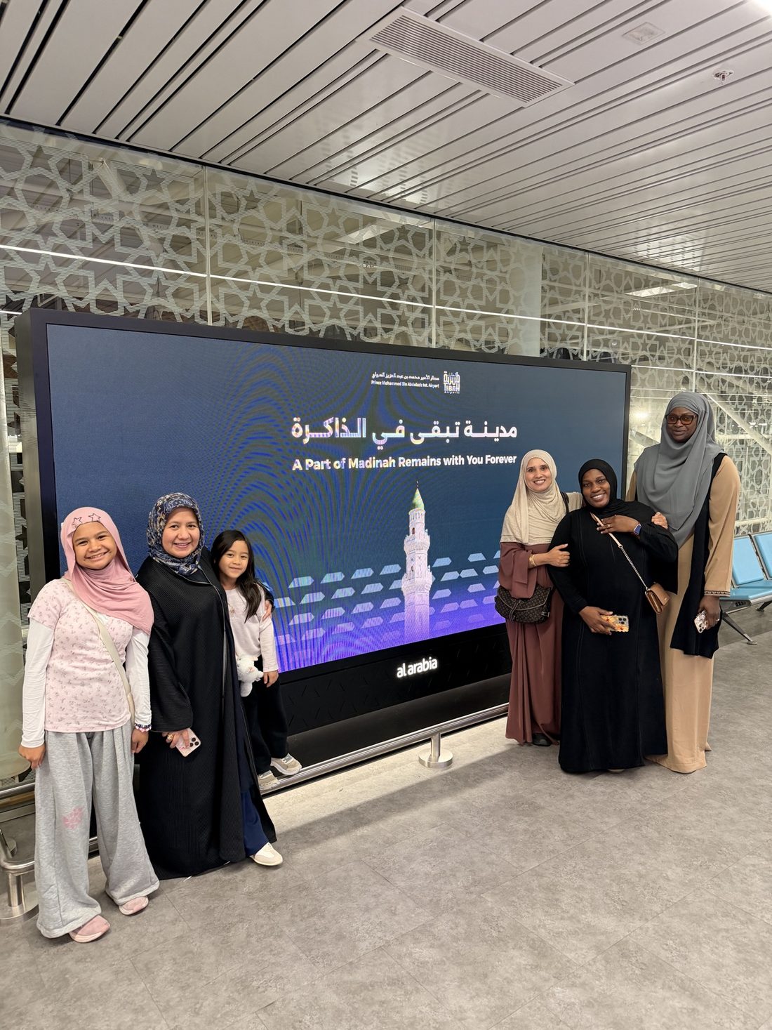 Members of the first Waseela group at Madinah airport beside a welcome screen reading 'A Part of Madinah Remains with You Forever'
