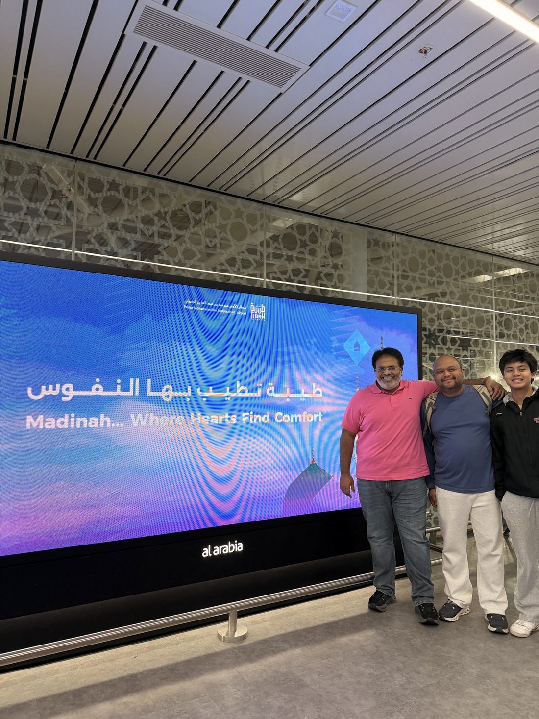 Three members of the group at Madinah airport in front of a screen reading 'Madinah… Where Hearts Find Comfort'